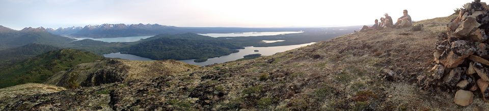 A panoramic photograph taken from the top of Hideout showcases both Hidden Lake and Skilak Lake. The Kenai Mountains are visible in the left-background of the image, and four hikers, resting in the alpine, are visible in the right-foreground.